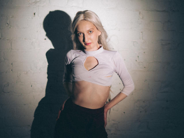 ViktoriaClark with long blonde hair, wearing a white crop top with a cutout, standing against a white brick wall, shadow on the wall, hands on her hips, looking at camera.