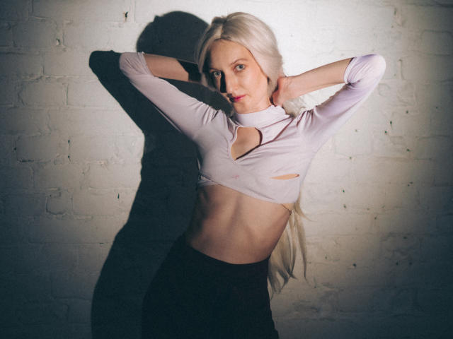 ViktoriaClark with long blonde hair, arms raised, wearing a light purple crop top with a cut-out, exposing her small tits and flat stomach, against a white brick wall.