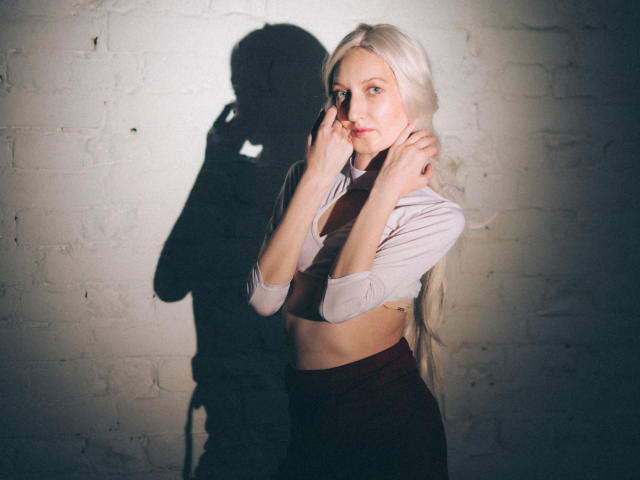 ViktoriaClark with long blonde hair, wearing a white crop top and dark skirt, touching her face, shadow on white brick wall, soft lighting, slightly parted lips, mid-shot.
