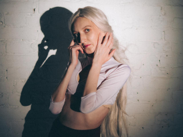 ViktoriaClark with long blonde hair, white shirt, black necktie, and black pants, hands on face, shadow on white brick wall, mid-shot, light skin, slim build, subtle smile.