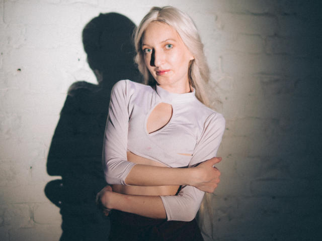 ViktoriaClark with long blonde hair, wearing a white crop top with a cutout, arms crossed, shadow on a white brick wall, looking at the camera with a slight smile.