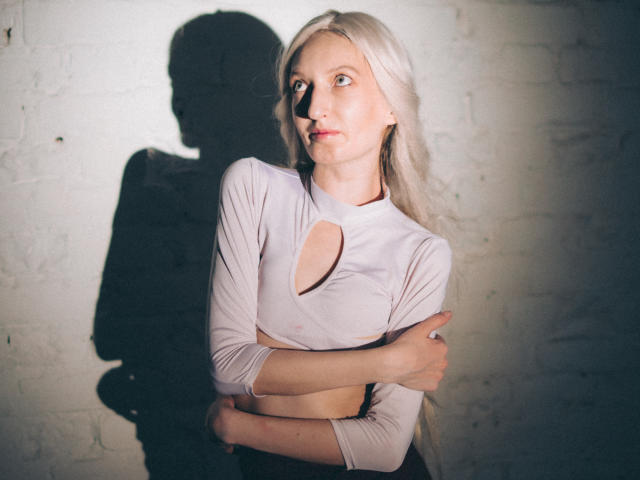 ViktoriaClark with long blonde hair, wearing a white cut-out top, arms crossed, shadow on white brick wall, looking to the side, soft light, serious expression.