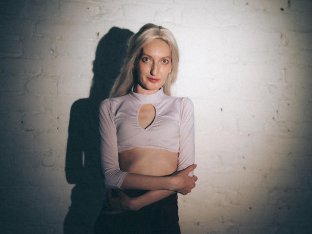 ViktoriaClark with blonde hair, pale skin, and small tits, wearing a sheer pink top with a cut-out, standing against a white brick wall, arms crossed, looking at camera.