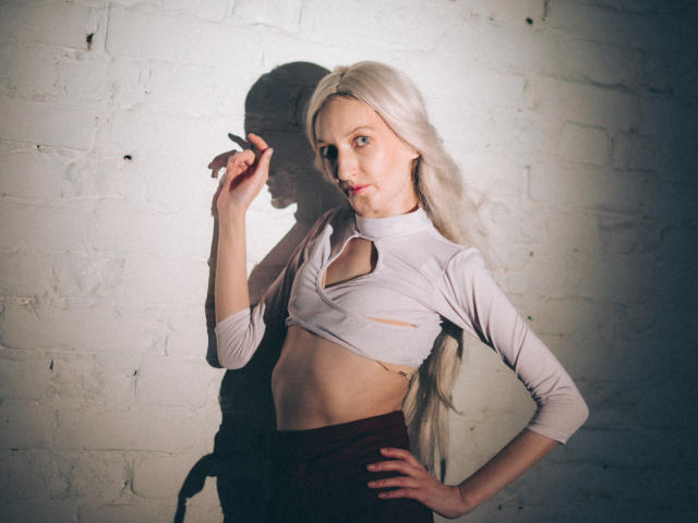 ViktoriaClark with long platinum blonde hair, white crop top, cut-out, exposing small tits, and maroon pants, hand on hip, finger near eye, standing against white brick wall.