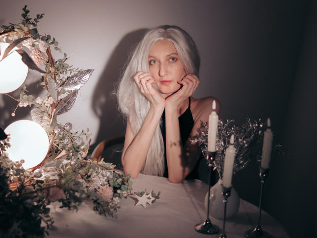 ViktoriaClark with long white hair, hands under chin, black sleeveless top, blue eyes, sitting at a table with candles and a decorated bouquet in a dimly lit room.