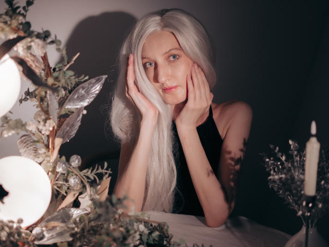 ViktoriaClark with long white hair, hands on cheeks, wearing a black sleeveless top, looking at camera, candle on right, leafy decoration on left, soft lighting, subtle smile.