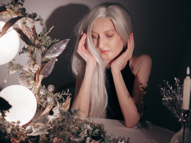 ViktoriaClark with long white hair, hands on her face, black sleeveless top, soft light, floral and candle decorations in foreground, gentle expression, subtle makeup, relaxed pose.