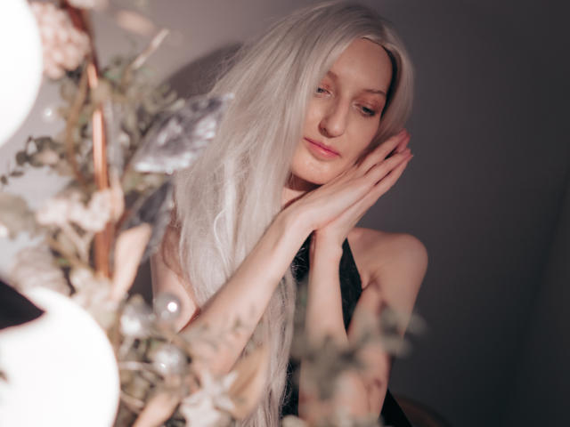 ViktoriaClark with long, white hair, black halter top, hands near face, soft smile, pink lips, delicate features, blurred floral decor in foreground, soft lighting, serene expression.