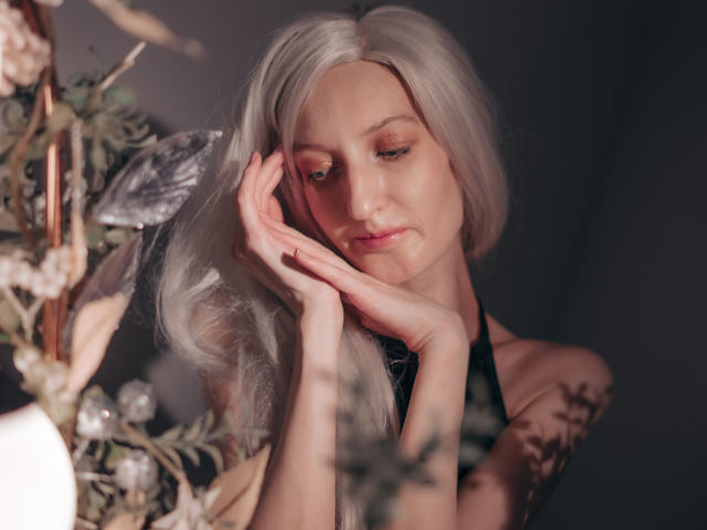 ViktoriaClark with long, white hair, touching her face, wearing a black halter top, showing tattoos on her right arm, soft lighting, and floral decor in the foreground.