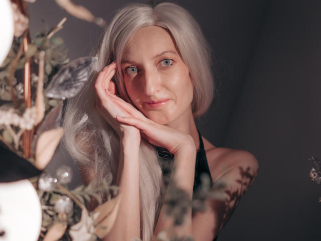 ViktoriaClark with long, platinum blonde hair, blue eyes, and a subtle smile, rests her chin on her hand, showing her delicate fingers and soft skin. Boobs not visible.
