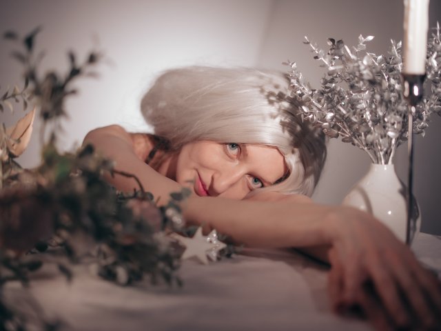 ViktoriaClark with platinum blonde hair, lying on a white surface, arms over face, surrounded by silver and green decorative branches, white candle in background, blue eyes gazing.