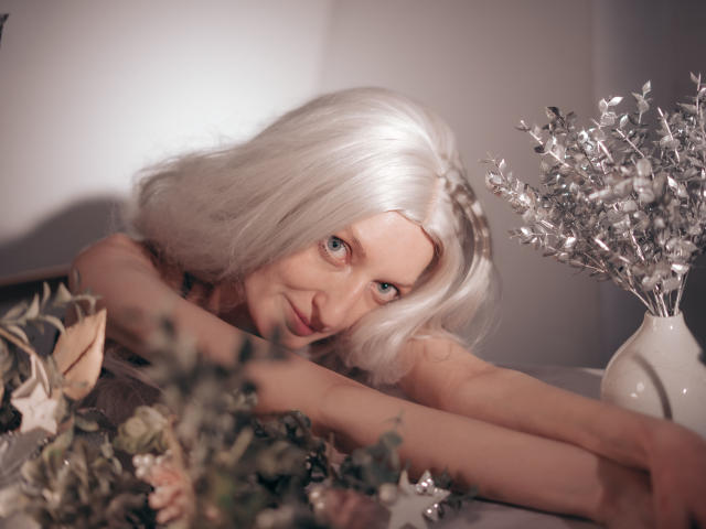 ViktoriaClark with silver hair, blue eyes, and a cute smile, lying down, arms crossed, surrounded by silver and green foliage, and a silver leaf vase on the right.