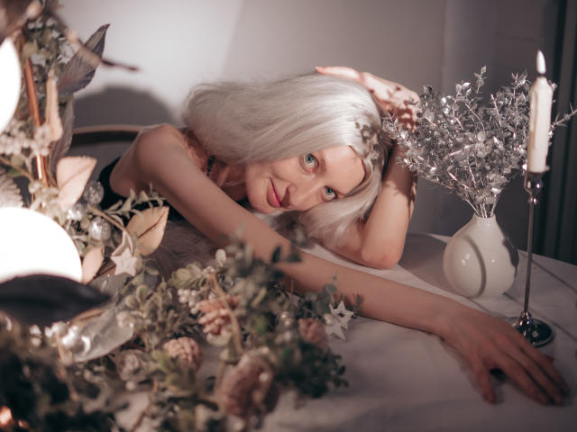 ViktoriaClark with white hair, lying on a table, arms stretched, looking at camera, candle, silver foliage, and flowers in background, soft lighting, sensual, calm expression.
