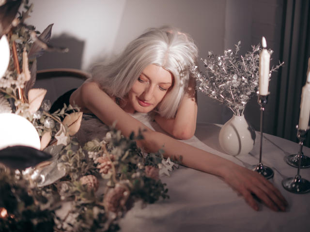 ViktoriaClark with white hair, lying on a table, arm stretched out, looking down, surrounded by candles, flowers, and a silver vase with white branches, in soft light.