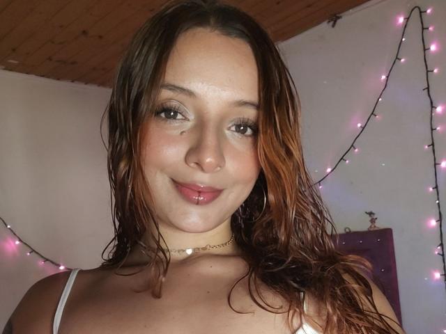 SaarahJones with wet brown hair, cute face, pink lips, white spaghetti strap top, choker, and pink fairy lights in the background. She's smiling softly at the camera.
