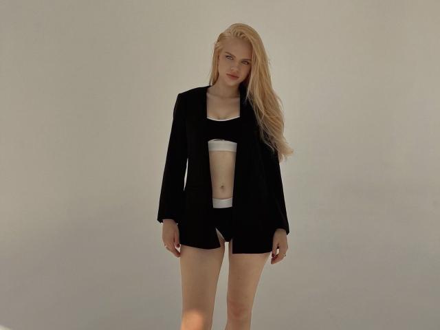 AviciAura with long blonde hair, black open-front shirt, black shorts, and white bra, standing, showing a small, flat-ass, and no visible tits, in a simple, plain background.