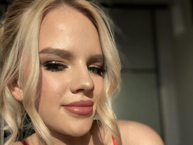 AviciAura has blonde hair, full lips with pinkish-nude lipstick, and dark, thick eyebrows. She's looking down with a slight smile, showing off her glossy, smooth skin.