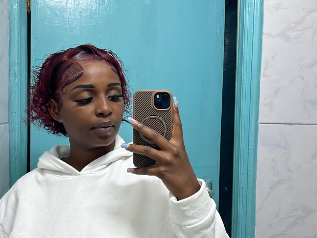 SteicyLovee, a dark-skinned woman with short, curly red hair, takes a mirror selfie. She wears a white hoodie and holds a phone with a brown, textured case. Her makeup includes subtle eyeliner and pink nail polish. She looks to the side with a calm expression. The turquoise door behind her contrasts with the white marble wall. The image is sharp with clear details. The composition focuses on SteicyLovee's face and upper body.