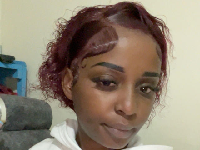 Portrait of SteicyLovee with dark brown skin and wavy, red-purple hair. She has thick, dark eyebrows with a noticeable gap in the right eyebrow, and subtle makeup with dark eyeliner. SteicyLovee is looking slightly to the right with a slight, confident smile. She is wearing a white hoodie. The image is a close-up, focusing on her face. The background is blurred, with a blue shelf and gray cushion partially visible. The lighting is soft, highlighting her smooth skin.