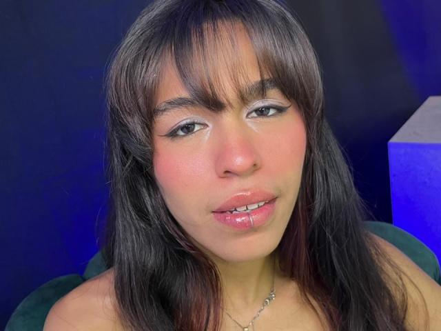 SoffiaCollins has straight black hair with bangs, shiny pink lips, and gray eye makeup. Her cute face is centered, with a blue light to the right.