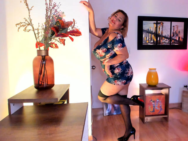 VanesaStella in floral dress, black thigh-highs, and black heels, leaning on door, curvy with big tits, red lips, smiling, wooden floor, vase with flowers, orange vase.