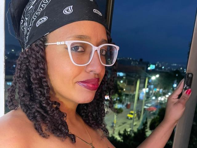 ArtemisaS has curly black hair, white glasses, red lipstick, and a black bandana, holding a phone, with a city night view in the background. Boobs not visible.