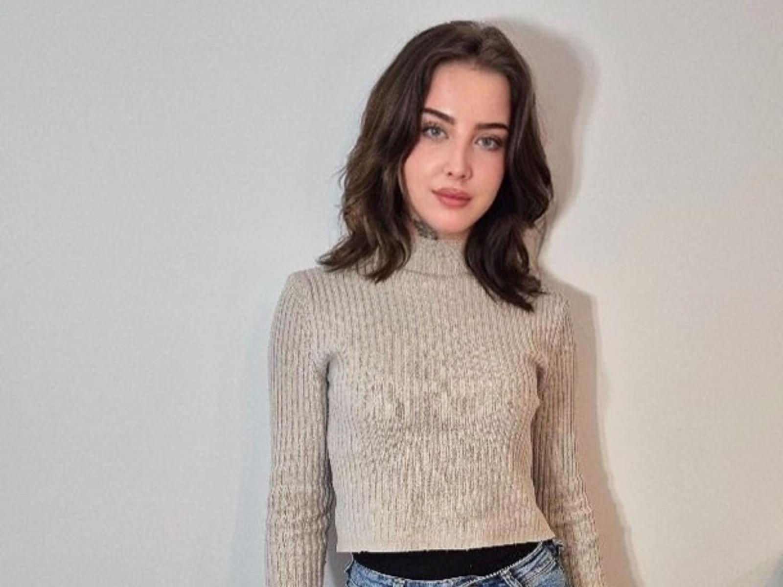 AmaraFlirt with wavy brown hair, wearing a beige rib-knit turtleneck, and black waistband, standing against a plain white wall, showing a subtle, confident smile.