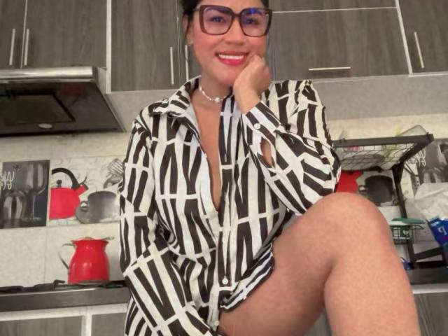 IvannaDuran, wearing a black and white striped shirt, sitting with one leg up, smiling with red lipstick, glasses, and a pearl necklace, in a modern kitchen.