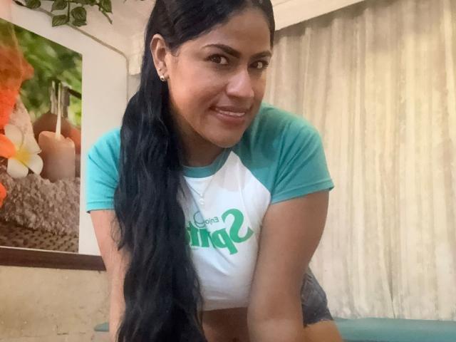 IvannaDuran with long black hair, smiling, wearing a green and white raglan shirt, leaning forward, showing her tits, in a room with a tropical painting.