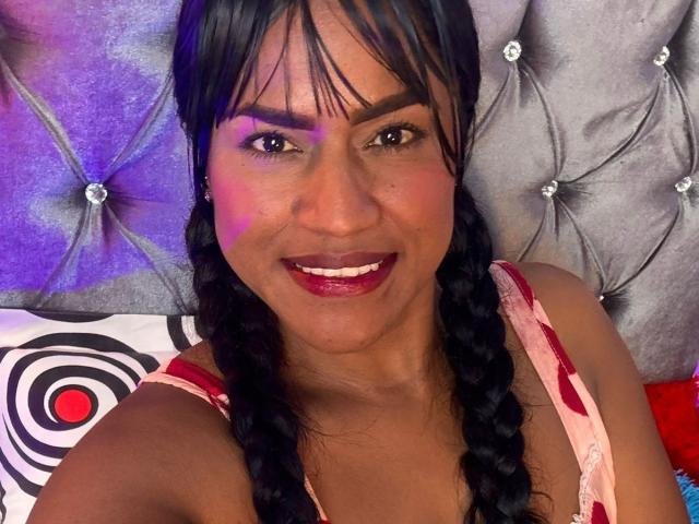 IvannaDuran with dark braids, red lipstick, and a smile, wearing a pink and red dress, has purple light on her cheek, sitting against a tufted, silver headboard.
