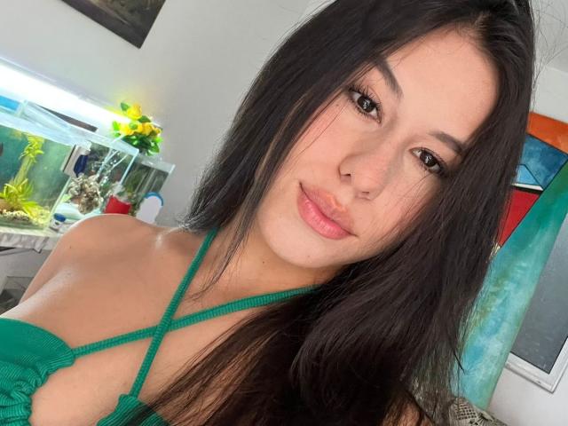 AmbarStoesselmml with long black hair, green strappy top, showing her tits, cute smile, brown eyes, and a nose piercing, in a bright room with a fish tank.
