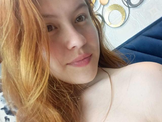 TaylorCarpert has red hair, fair skin, and is smiling with her pink lips slightly parted. Her bare shoulders are visible, showing a soft, natural look.