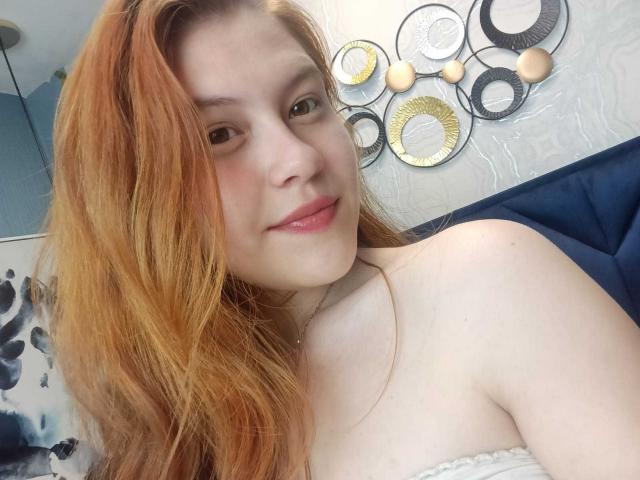 TaylorCarpert has light skin, orange hair, and is smiling with pink lips. She's topless, showing her bare shoulders. Her tits are small, and she's wearing a necklace.