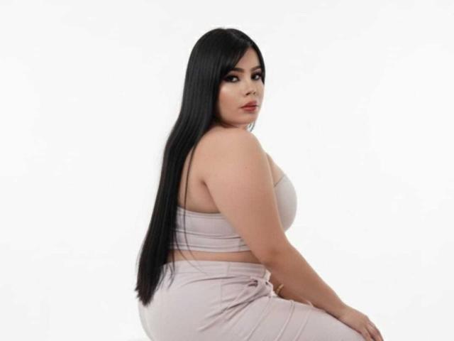 NathalyRose69 has long black hair, pale skin, and wears a pink strapless top with matching pink pants, showing her big ass and tits. She's looking back with a serious face.