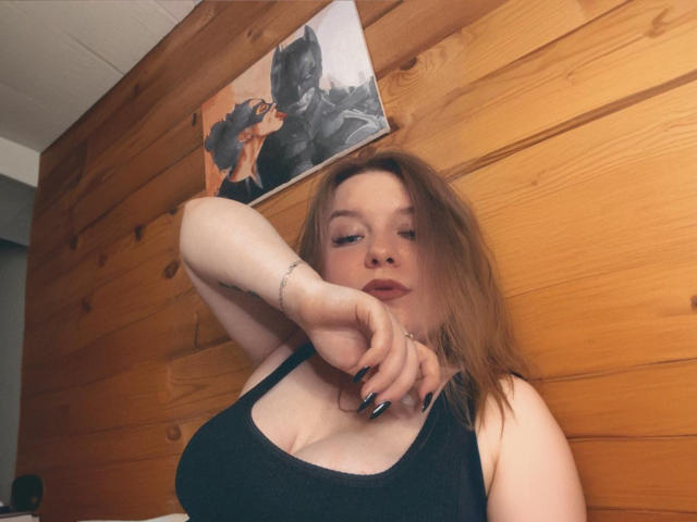 TwixyXRose with pale skin, red hair, and black tank top, showing off her large boobs and black nail polish, arm raised, looking at camera, wooden wall behind.