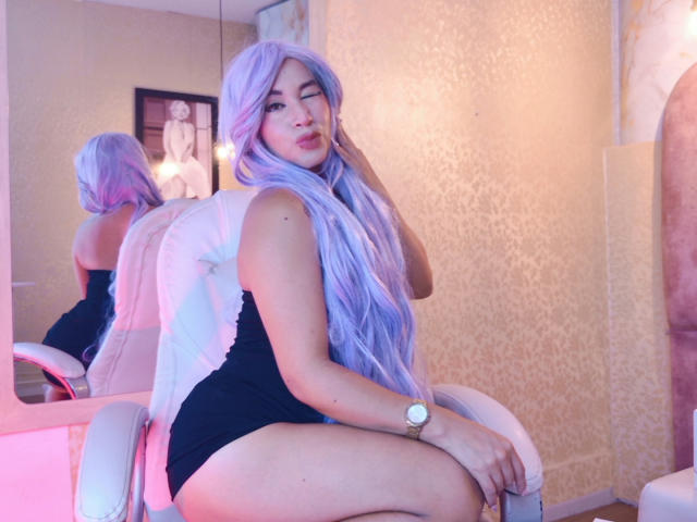 MissKendal with long, purple wig, black and white bodysuit, sitting on white chair, butt visible, wristwatch on, winking, pink and blue lighting, reflection in mirror.