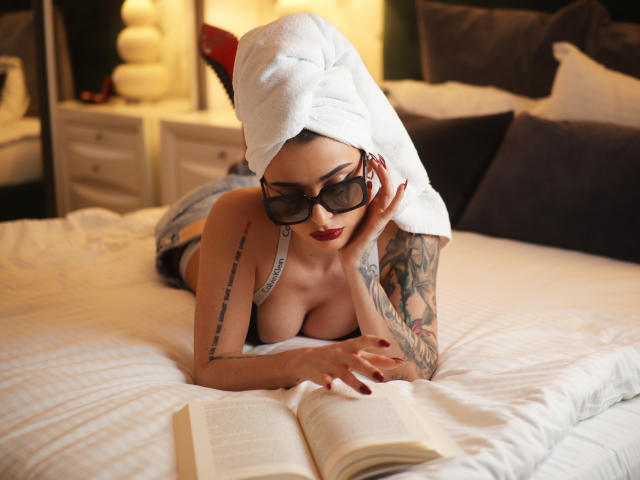 Venusinylons, avec une serviette sur la tête, portant des lunettes de soleil, allongée sur un lit, lisant un livre, montrant ses tatouages et ses seins dans un soutien-gorge blanc, avec des ongles rouges..