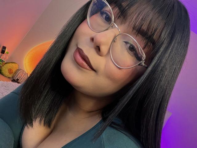 Close-up of AlexaKiim with straight black hair, wearing large clear glasses and dark lipstick. She has a light skin tone, full lips, and a slight smile. She's wearing a dark green top with a low neckline, showing a hint of cleavage. The background has a purple and orange gradient, with a small bottle and decorative object on a surface to the left. The lighting is soft, highlighting her face and glasses. Her bangs are straight and frame her face.