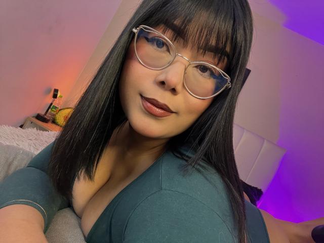 Close-up of AlexaKiim with straight black hair and bangs, wearing large, clear glasses and dark lipstick. She's looking at the camera with a slight smile. She has light brown skin and is dressed in a teal, low-cut top that shows cleavage. The background has a soft, pinkish-purple light. Her glasses reflect light, adding a shiny effect. The overall composition is centered on her face and upper torso. The image has a modern, casual style.