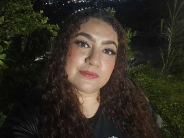 SeleneFhox6 is a medium-skinned woman with curly dark brown hair wearing a black shirt. She has thick eyebrows, long eyelashes, and red lipstick. Her expression is neutral with a slight smile. The background is dark with green foliage on the left and right. The image is a close-up portrait with natural lighting. She looks directly at the camera.