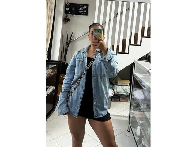 Velka with a camera in hand, wearing a light blue denim shirt, black shorts, and a "LADY BOSS" strap, standing in a room with a staircase.