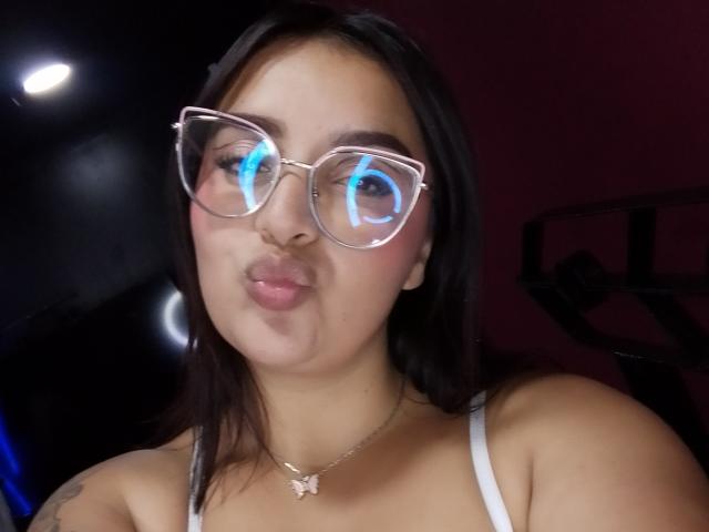 MissSubmissive hat braunes Haar, klare Brillen mit blauer Lichtreflexion, volle Lippen und ein weißes Tanktop, das ihre kleinen Brüste und eine Schmetterlingskette zeigt..