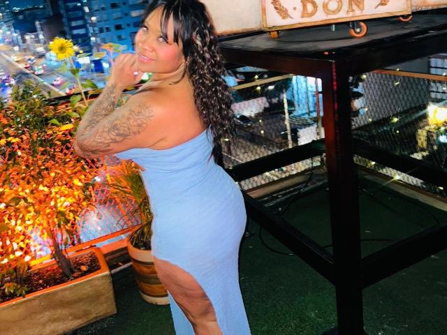 CloemaraDuval with tattoos on her arm, wearing a blue, strapless, high-slit dress, smiling, standing by a plant, night city lights in the background, curly brown hair.