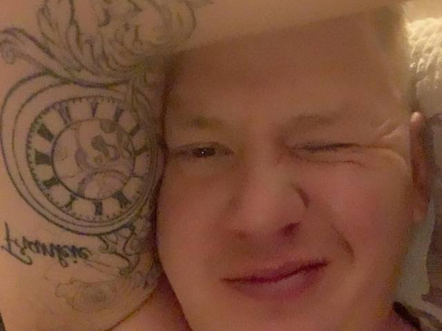 RemyBartss has a chubby face, one eye closed, smiling with a tattoo of a clock on their arm. They have a nose piercing and are in a dimly lit photo.