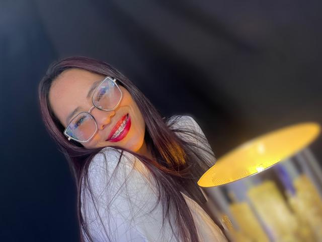 SiennaMoretti with long brown hair, glasses, red lipstick, smiling, white top, yellow lamp in background, slightly tilted head, cheerful expression, soft light.