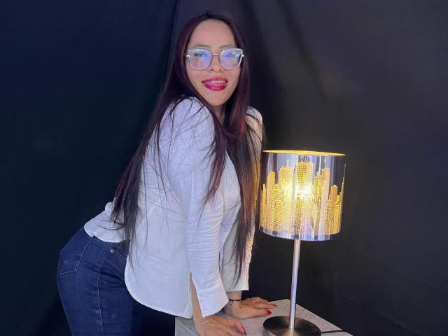 SiennaMoretti with long dark hair, glasses, red lipstick, white top, blue jeans, leaning on table, smiling, hands on table, next to a city-lights lamp.