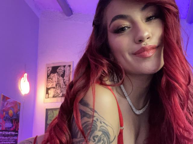 EvaMendex with red hair, nose ring, and lip gloss, wearing a red strap, necklace, and tattooed arm, smiling at camera, purple and blue lighting. Boobs visible, cleavage.