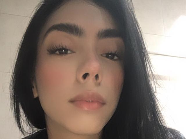 Alle69 has a cute face with dark eyebrows, long black hair, and glossy pink lips. Her nose is small and she's looking slightly to the side.