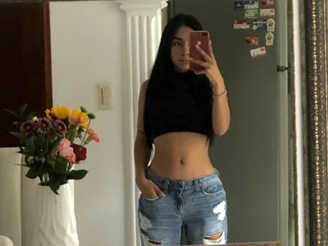 Alle69 with long black hair, wearing a black crop top, showing a flat, toned midriff, and ripped blue jeans, taking a mirror selfie with a red phone.