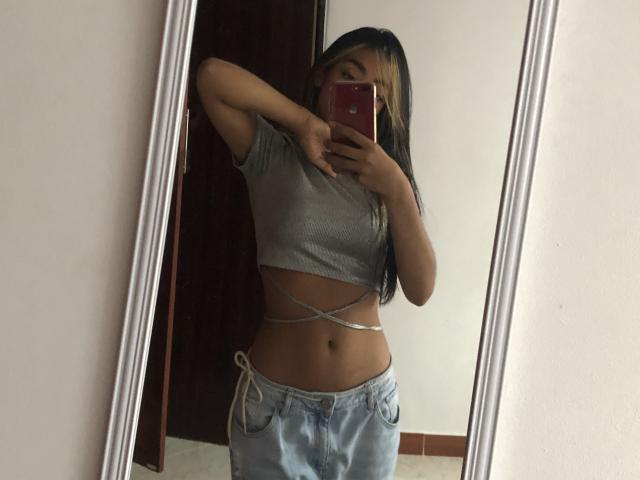 Alle69 with medium brown skin, long black hair, small tits, wearing a gray crop top, blue jeans, and a silver belly chain, taking a mirror selfie with a red phone.